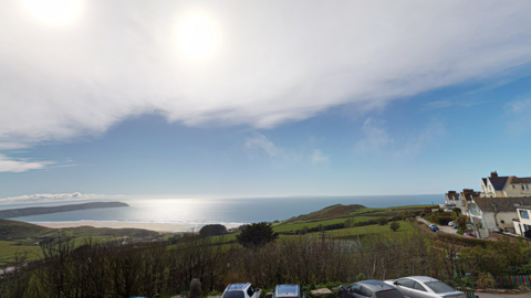 A scenic coastal photo looking out from a hotel balcony with the sea in the distance. Bright blue skies with the blue sea below. There is lots of green fields as well as some buildings to the right hand side of the photo. There are also a few cars parked and a sandy beach in the background.