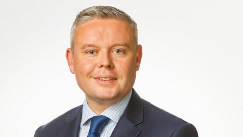 Keith Haslett  is smiling at the camera. He has short grey hair and is wearing a blue suit and tie