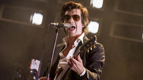 Alex Turner performs live. He's playing the guitar, wearing sunglasses and has a dark jacket on.