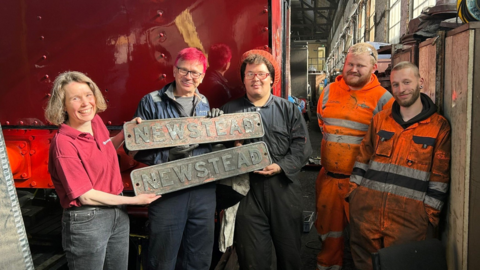 Two name plates are held up by three people standing in front of a red train, two men in orange hi-vis clothing stand to the right of the picture. Everyone pictured is smiling.