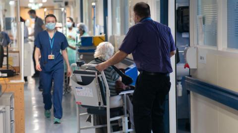 A hospital corridor. A man holds the back of a seat with an elderly patient in which white hair. Another member of staff in blue scubs and a blue face mask walks down the corridor. 