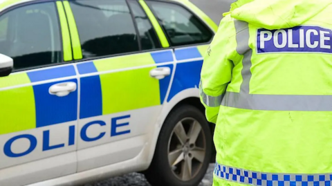 A police officer wearing a hi-vis jacket is pictured from behind. They are looking towards a marked police car. 