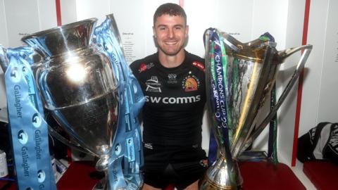 Ollie Devoto sat in a changing room wearing Exeter Chiefs kit in 2020 with the Premiership trophy on one side and Champions Cup trophy the other