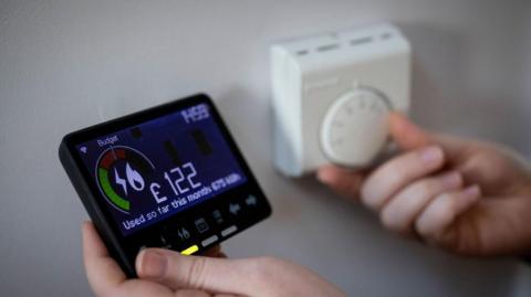 A pair of hands - one hand is holding a smart meter showing the money spent on electricity and another is adjusting a thermostat.