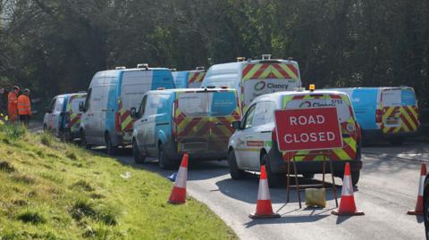 Several corporate vans are parked, bunched up on a road, as workmen work in the distance. There is a red road closure sign and a number of orange road cones. 