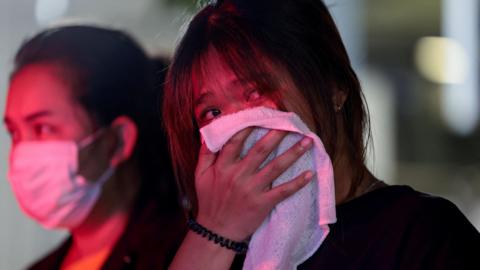 Two women stand, their faces lit by emergency services lights, one wearing a face mask and the other holding a white cloth over her mouth and nose. 