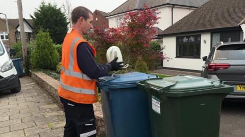 A refuse collection worker in hi-vis clothing picks up a kettle left out on a blue recycling bin from a residential street. 