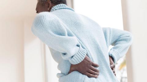 A woman wearing a pale blue jumper is seen from the back. She is leaning forward and has her hands placed on her lower back.