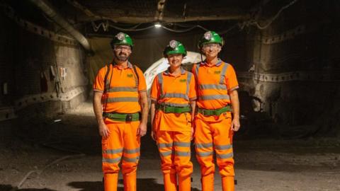 Two men and a woman in orange hi-vis gear and green belts with green helmets stand in the STFC Boulby Underground Laboratory