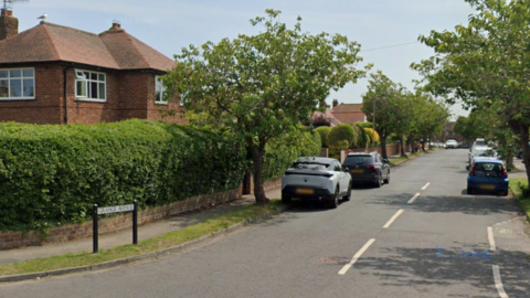 A quiet residential street with trees along the verge and cars parked up