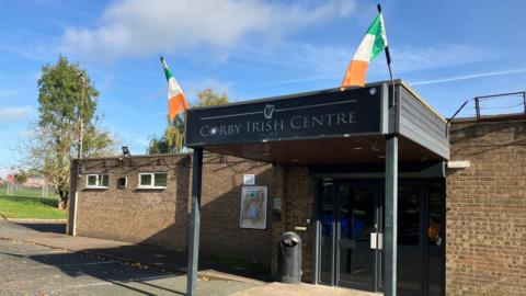 A square brick building set next to a green bank with trees, with a concrete car park in front. The building has a black wooden awning with pillars at the front and displays a black sign saying 'Corby Irish Centre' in silver lettering. At the two front corners there are Irish flags, in orange, white and green colours. There is a blue sky in the background.