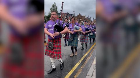Bagpipers wearing purple top and a variety of red and green kilts, walking down a concrete road with double yellow lines. The street is in Glasgow. There are houses in the background and the sky is grey and cloudy.