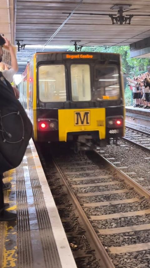 Yellow metro train arriving at a station platform, with people standing nearby and a group on the opposite platform waving toward the train.