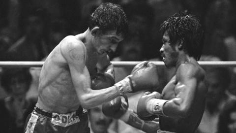 A black and white image of two men wearing shorts and gloves boxing in a ring 