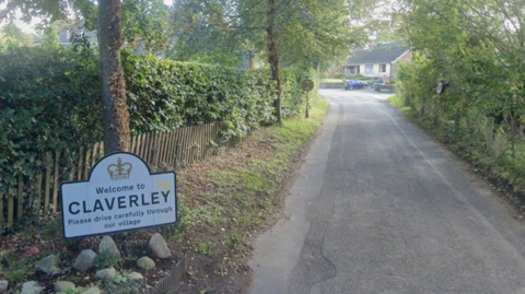 A country road with a thick hedgerow on the left side and a thin line of trees to the right. There is also a white road sign in the foreground for Claverley.