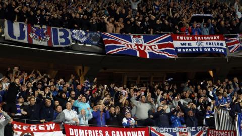 Rangers fans at the match with Nice, many dressed in replica kits. Many of the supporters are chanting and singing, while banners are draped around their section listing which area they are from. 