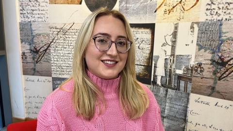 Woman in pink jumper with long blonde hair and glasses, pictured against a wall of archive letters 