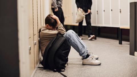 A child sitting on the floor next to some lockers. They have a black bag next to them