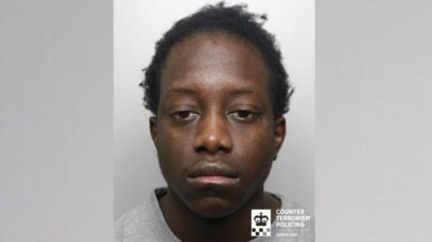 A mugshot of a young man with short black hair and wearing a grey jumper.