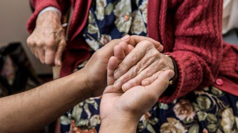 The hand of an elderly woman in in a red cardigan and flowery dress is being held in both hands by a care worker. Their faces are not visible.
