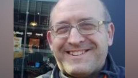 A man wearing glasses and smiling, he is standing in front of what looks like a shop window.