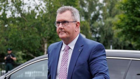 Jeffrey Donaldson is walking away from a black car.
He is a man with short grey hair, wearing a dark blue suit, white shirt and pink tie.