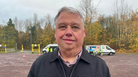 Richard pictured smiling slightly and looking into the camera. He has grey hair. He's wearing a dark grey zip-up jacket, with a red and blue checked shirt and dark grey woollen jumper on underneath. He's stood in a muddy gravel car park, which is surrounded by trees which have orange and dark green leaves. Beyond him, at the edge of the car park, are two vans and a police motorbike. The sky is grey and overcast. 