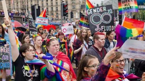 A group of people dressed in rainbow flags carrying banners walking through Liverpool city centre.