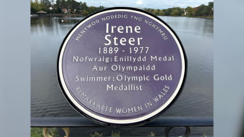 A purple plaque with silver writing and trimming. The plaque says 'Menywod nodedig yng Nghymru'. Irene Steer, 1889-1977. Nofwraig: Enillydd Medal, Aur Olympaidd, Swimmer: Olympic Gold Medallist. Remarkable women in Wales'. 