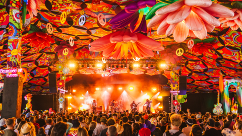 A stage at Kendal Calling. A four-piece band is performing on stage to a crowd of hundreds of people. The inside of the tent is decorated with brightly-coloured flowers and peace signs to give it the feel of a 1960s hippy experience.