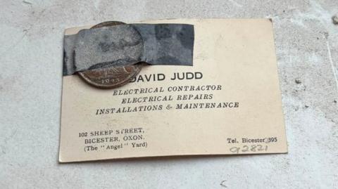 A coin is taped to a business card which bears the name "David Judd, electrical contractor"