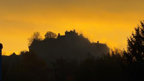 A dark outline of a hilltop castle against a glowing orange sunrise sky.