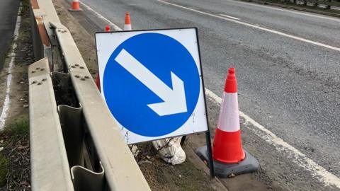 A 'keep right' white arrow in a blue circle on a road sign stands between a central reservation and an orange and white traffic cone. There are other traffic cones in a line behind the sign, on the dual carriageway.