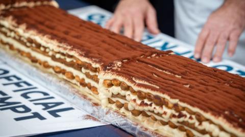 A very long tiramisu on a table. There are a person's hands behind it