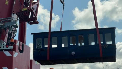 A blue train carriage is being lifted up into a sunny sky by a red crane.