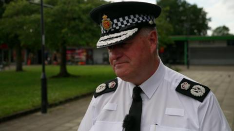 A man with short grey hair is wearing a white shirt with chief constable on black lapels, along with a black tie and a chief constable's peaked cap. He is being interviewed while standing on a paved area with a shop, trees and a grassed area behind him.