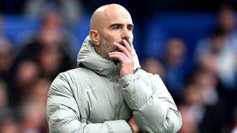 Enzo Maresca, looks on during the Premier League match between Chelsea and Brighton at Stamford Bridge. He is wearing a grey coat and holding his face.