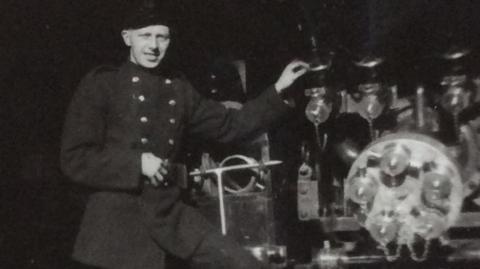 A black and white picture of William Crocket standing on a piece of machinery. He is wearing a dark jacket with buttons on either side.