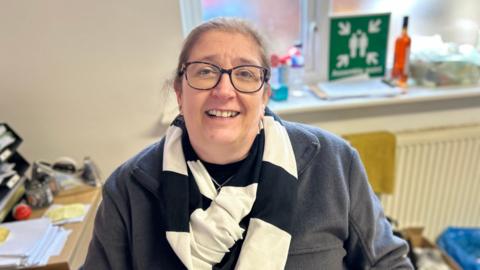 A lady looking up at the camera. She has glasses and a black and white scarf on. She is smiling.