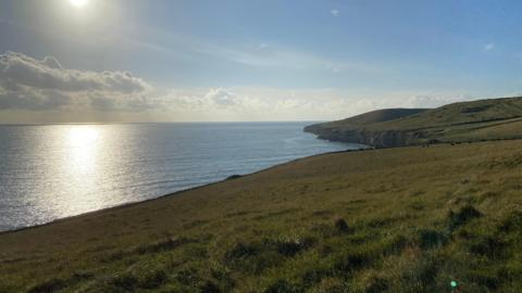 A stunning beach scene with a grassy landscape looking out over the sea. The sun is low in the sky which is clear and blue apart from some light white clouds.