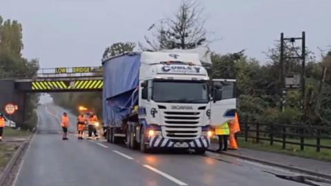 Crashed lorry with emergency service workers around it and bridge in background