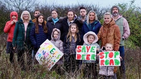 A group of 14 people opposing the plans standing in a field. Three of the four children standing in front of the group of adults are holding signs reading Trees Not Trucks, Save Our Deer and Stop The Road.