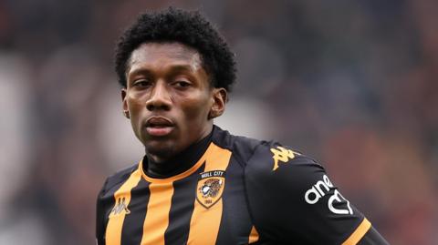 A close up head and shoulders picture of Jaden Philogene, wearing Hull's black and amber striped kit looks across the camera and slightly to his left