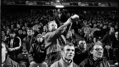 A close-up, shot in black-and white, of a football fan standing up and pointing to his watch while other supporters around him remain seated. 