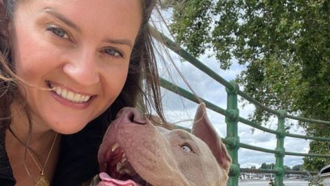 Sophie Coulthard, a woman with long brown hair wearing a black top and gold necklace, posing with her dog, a grey XL Bully, in front of green railings