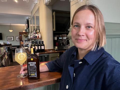 A woman with blond hair and a blue shirt stands in front of a bar with a brown bottle of Mother Root and a drink in a wine glass