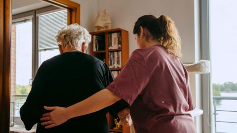 An elderly woman with short white hair is escorted by a woman with long brown, wearing maroon scrubs. The younger of the two has her left hand on the woman's back. The elderly woman wears a black dress. A wooden book shelf, two shelves wide and four long is partially stacked with books. There are two windows either side of the bookshelf, a body of water is visible outside.