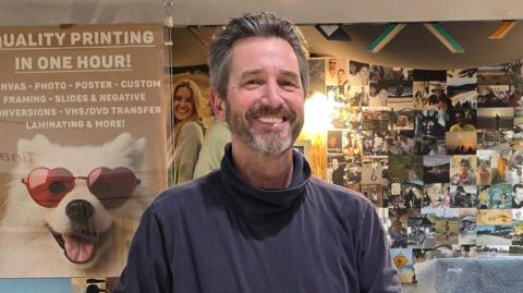 Simon Stockdale smiles as he stands in front of the glass window of his print shop. He has grey-brown swept back hair and a beard.