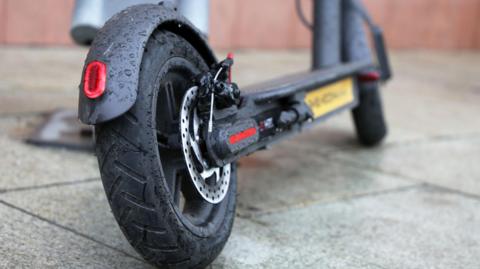 A close up of an e-scooter's wheels. It is a black bike with black tires and is standing on a footpath.