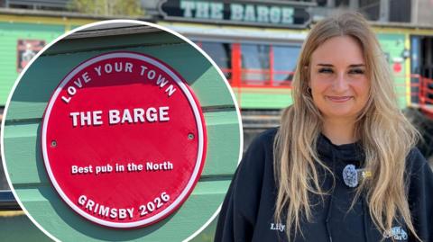 Woman with long blonde hair smiles to camera. She is wearing a dark, navy jacket with her name 'Lily' embroidered into it. In the background The Barge, a bright mint green boat with red detailing around the glass windows can be seen. There is also a close-up picture of the red plaque which reads: 'Love Your Town, The Barge, Best pub in the North, Grimsby 2026'.